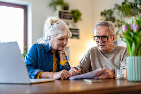 An elderly man and woman sit at a wooden table, reviewing papers together. A laptop and a vase of pink tulips are also on the table.