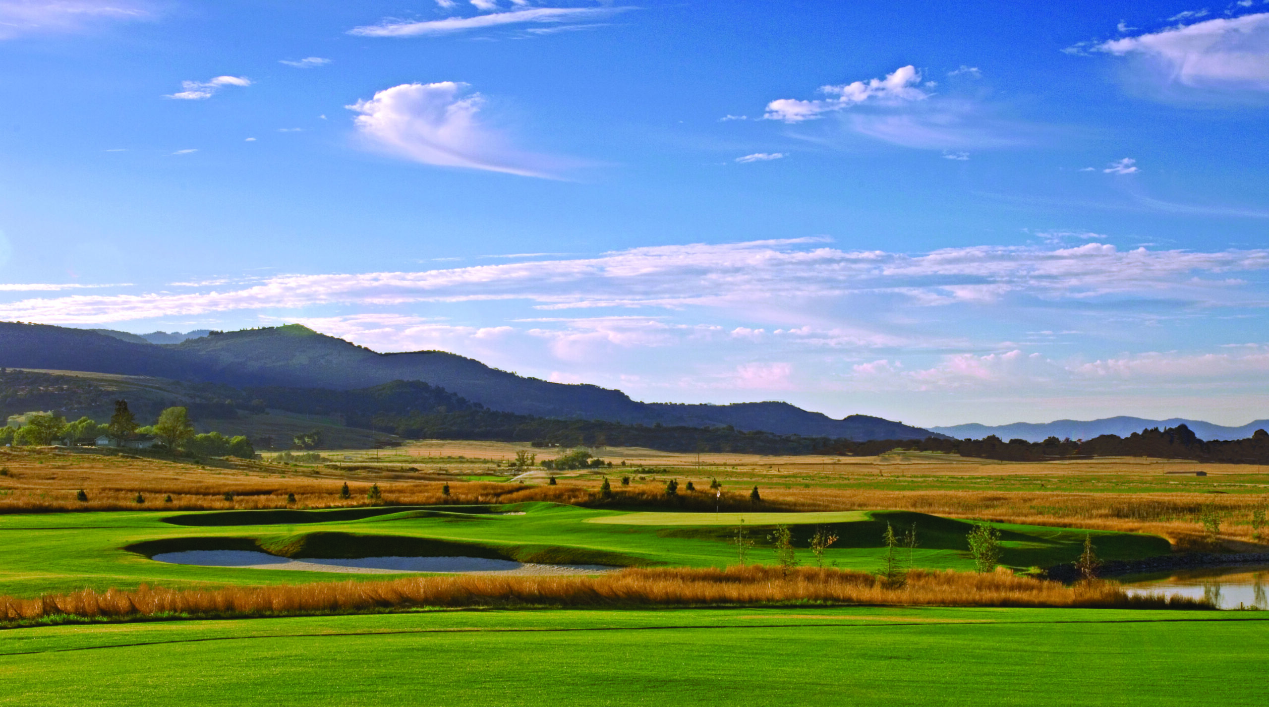 A panoramic view of a golf course with green fairways, sand bunkers, a pond, and golden grasses, set against a backdrop of dark mountains under a blue sky with white clouds.
