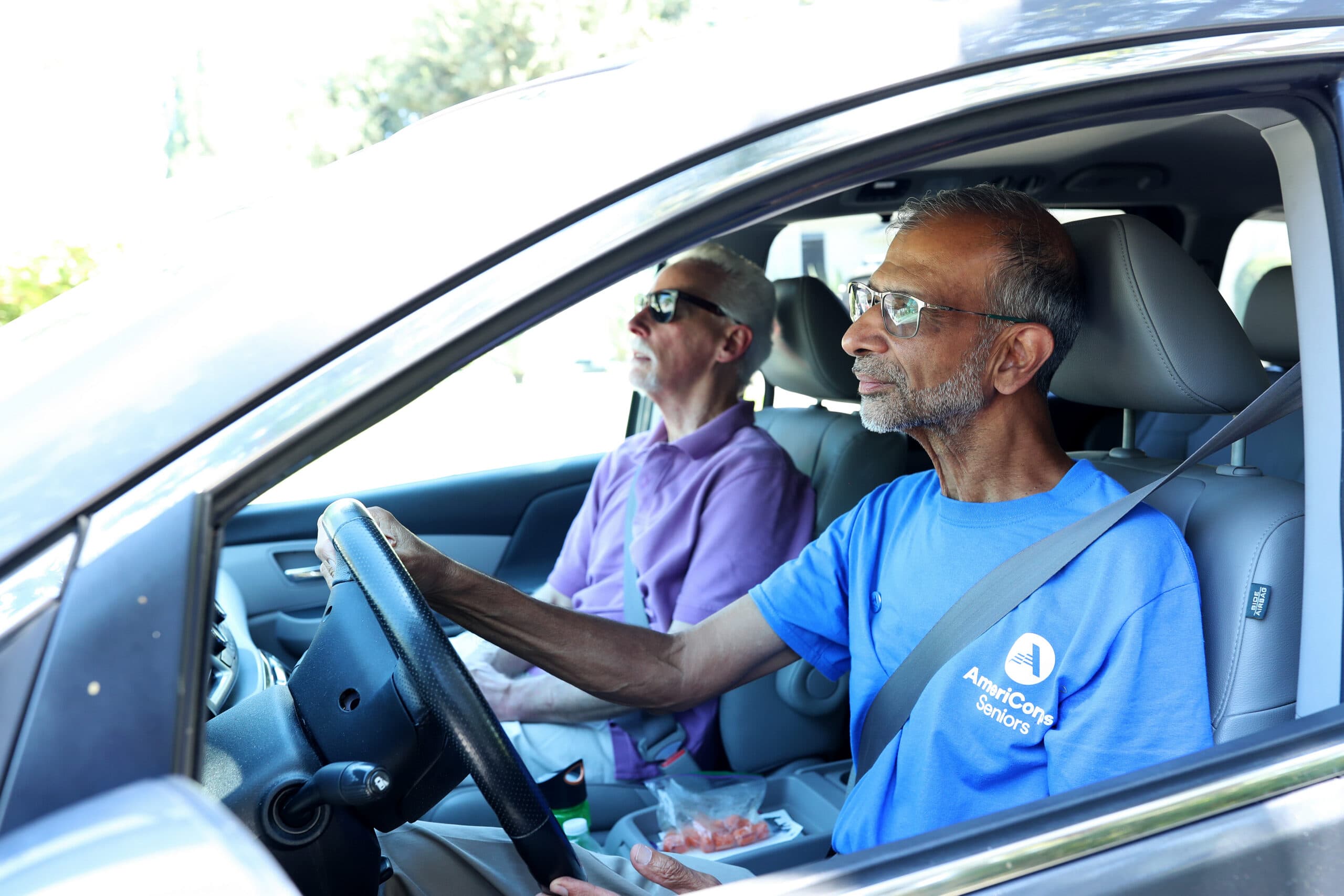 Two older men in a car. The driver wears a blue AmeriCorps Seniors shirt and glasses, while the passenger wears a purple polo and sunglasses.