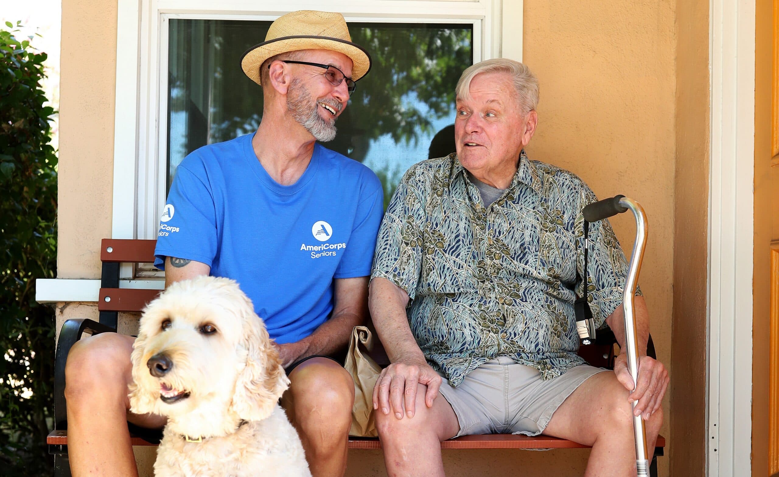 Two men and a curly-haired dog sit on a bench. One man wears an AmeriCorps Seniors shirt and a fedora, smiling at the other man who holds a cane. The dog sits between them.